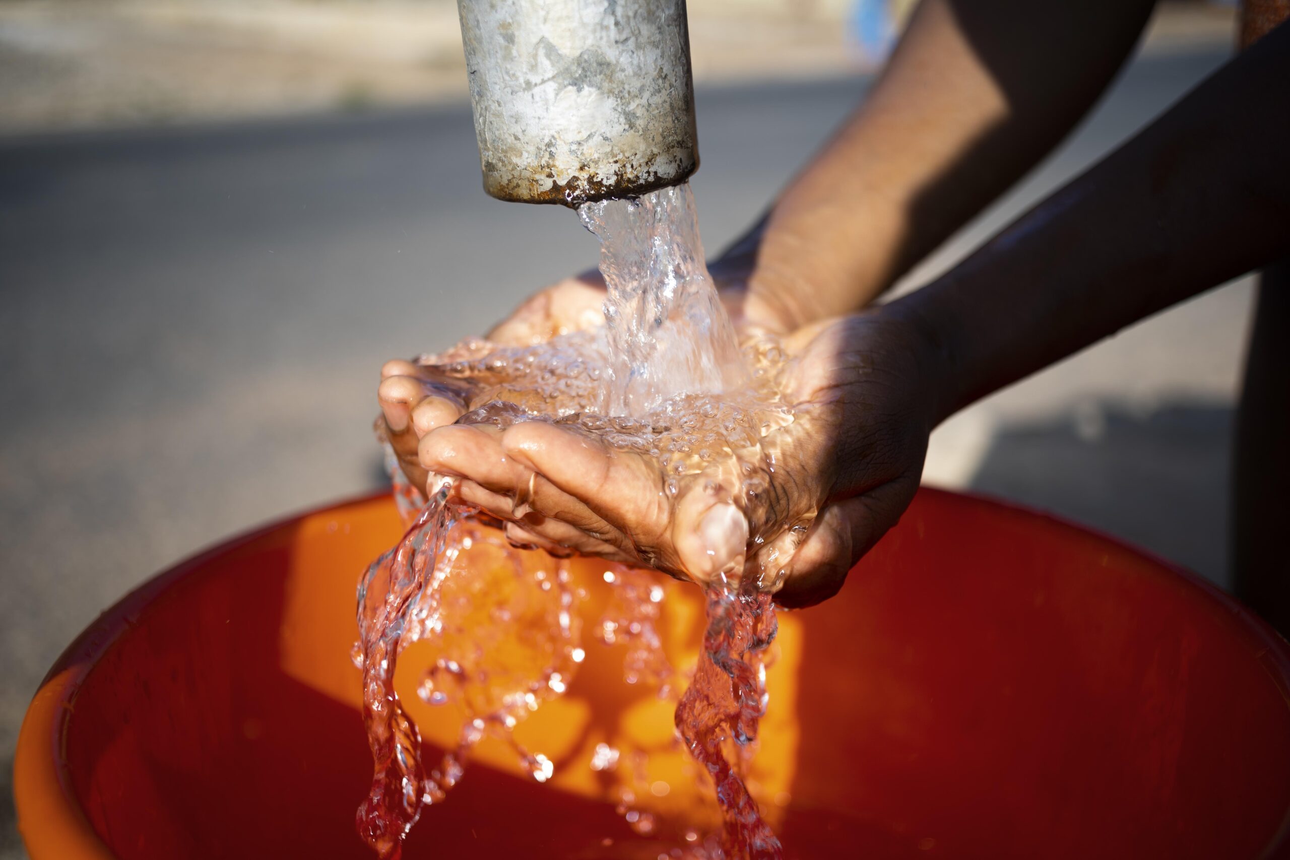 Manos recogiendo agua limpia de una tubería, con un recipiente naranja colocado debajo.