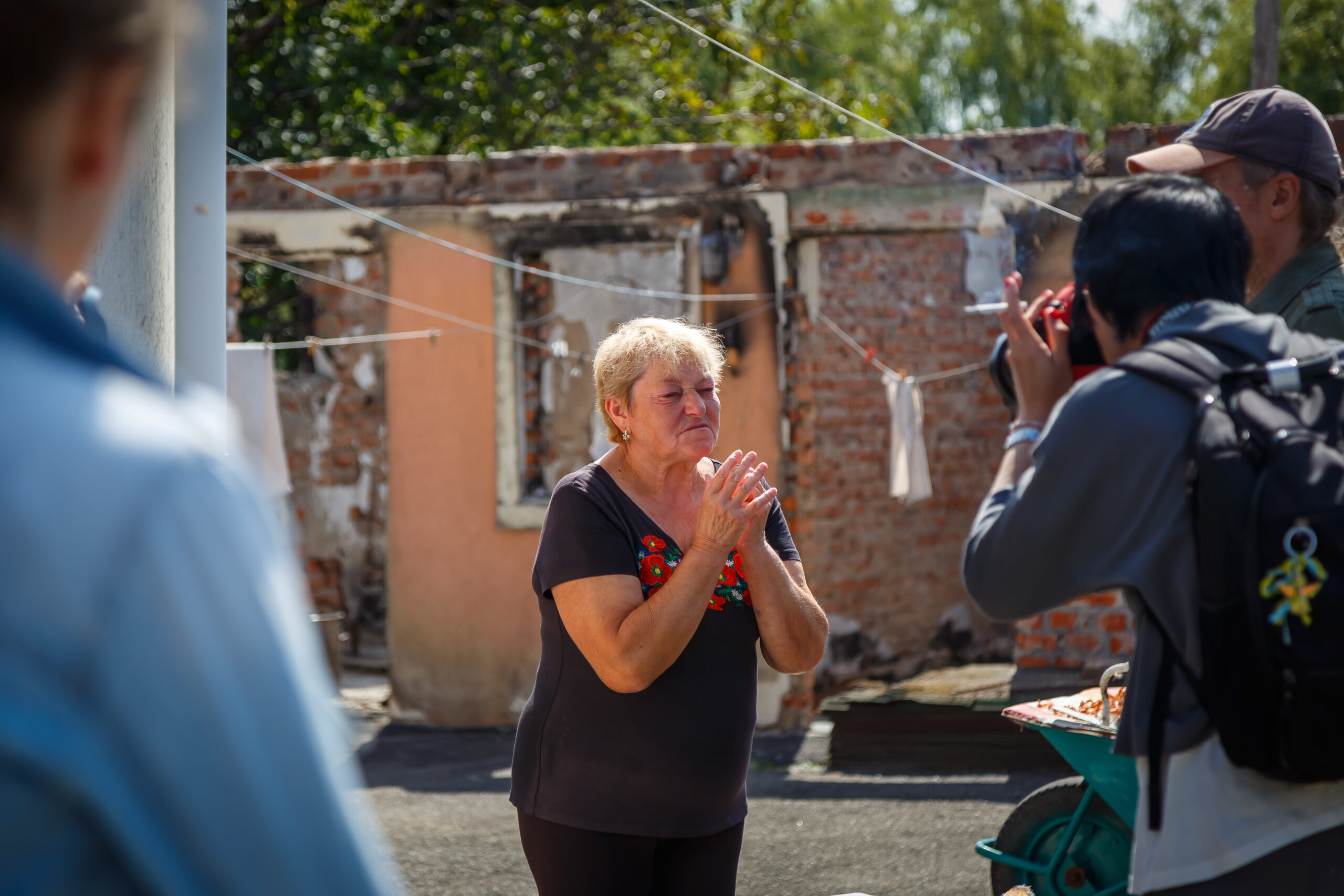 Mujer mayor emocionada ante los restos de una casa destruida mientras un fotógrafo documenta la escena