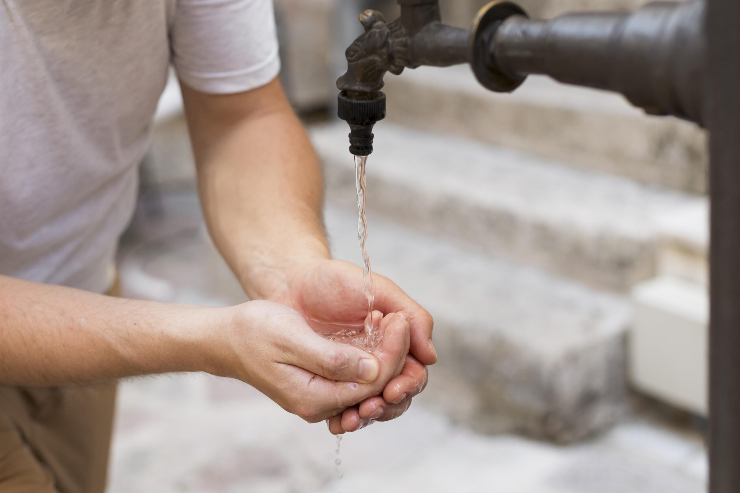 Persona juntando agua con las manos bajo una canilla de metal en un entorno urbano.